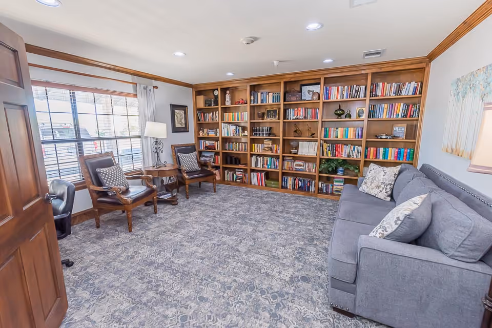 Cozy sitting room with large wooden bookshelves, two armchairs by a window and a gray sofa on a patterned carpet.