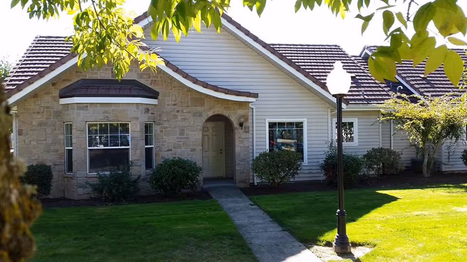 Exterior view of a single-story residential building with a stone and siding facade, a tiled roof, a front door under an archway, windows, bushes, a green lawn, a concrete walkway, and a black lamp post. Tree branches with green leaves frame the top of the image.