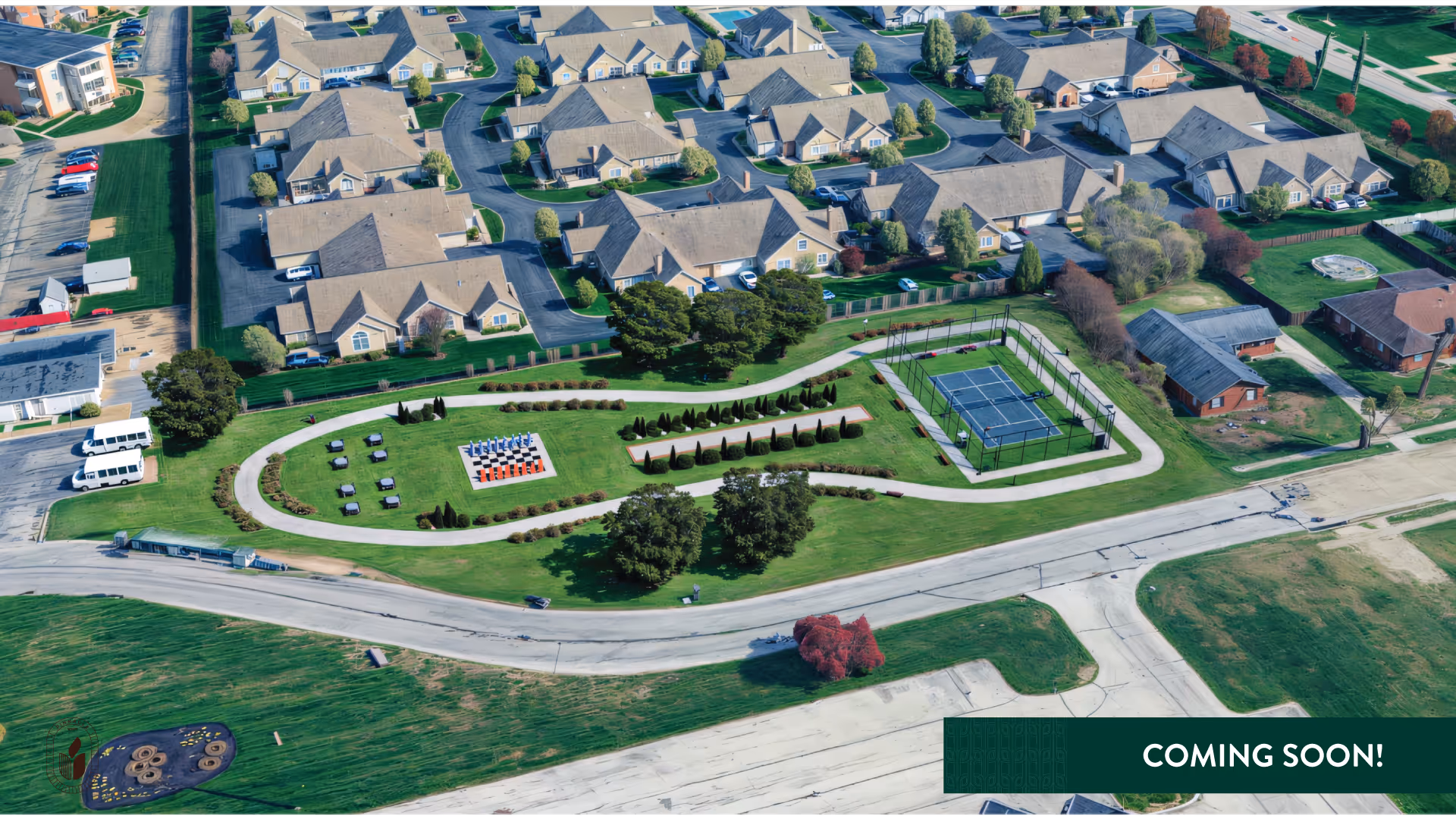 Aerial view of a senior living community showing clustered cottages, landscaped lawns, walking paths, outdoor seating, and a fenced tennis/pickleball court.