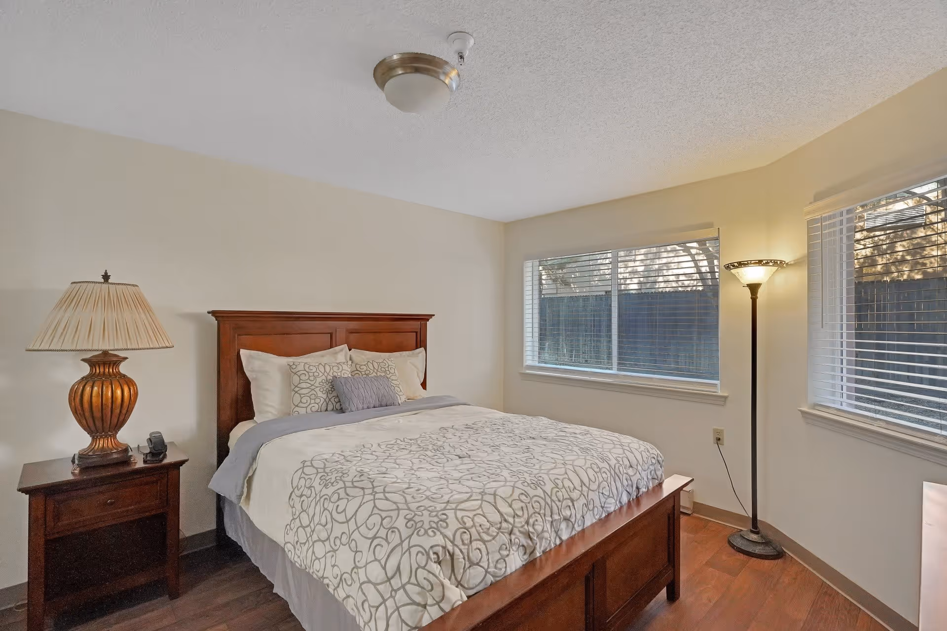 A bedroom with a wooden bed frame and a neatly made bed with patterned bedding. There is a wooden nightstand with a decorative lamp and a phone on it to the left of the bed. Two windows with blinds allow natural light into the room. A tall floor lamp is positioned in the corner near the windows. The walls are painted light beige and the floor is wooden.