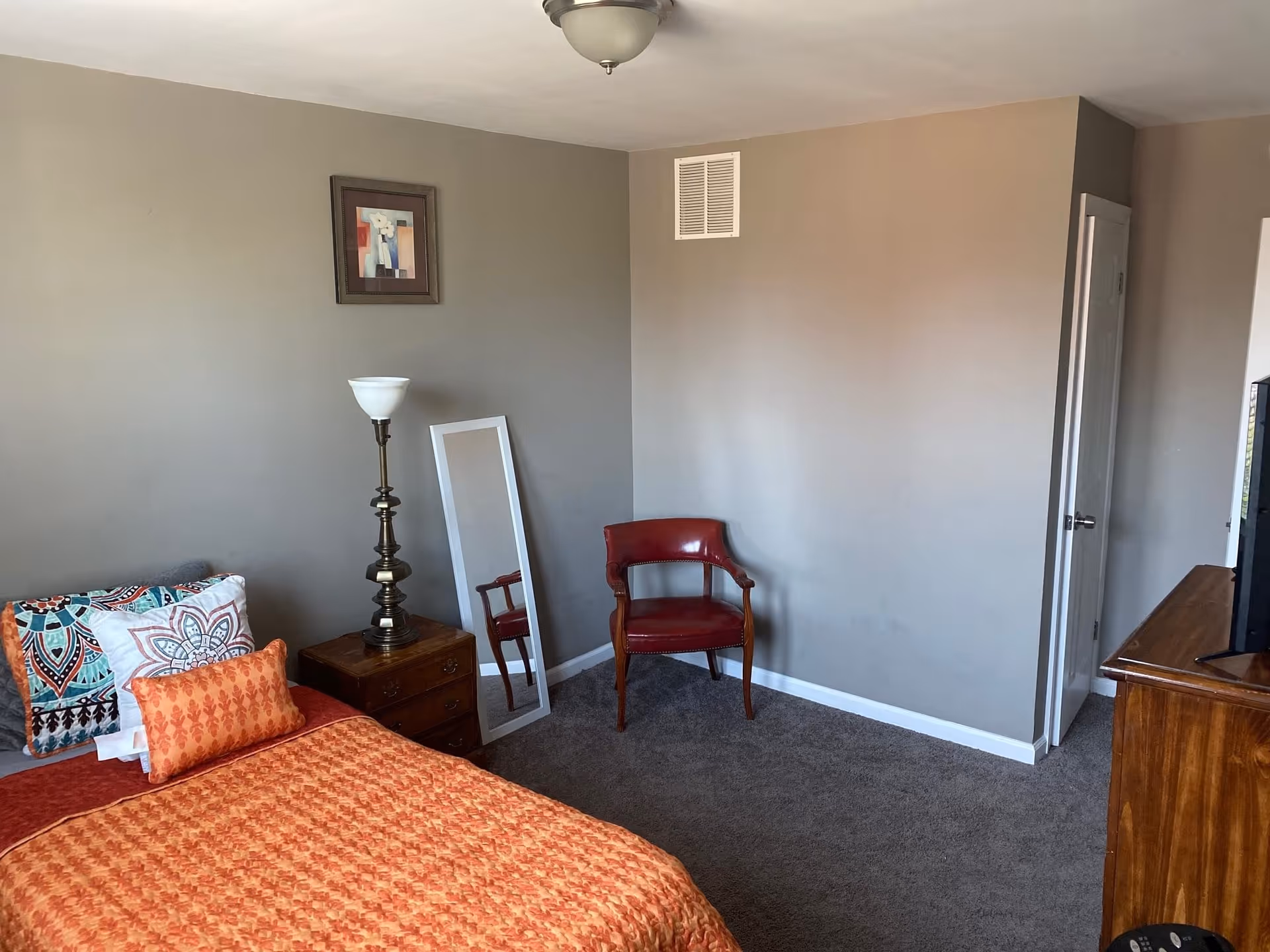 A bedroom with an orange bedspread, patterned pillows, a bedside table and lamp, a full-length mirror, a red chair, and a wooden dresser.