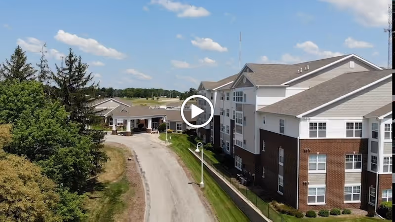 Aerial view of a multi-story senior living facility building with a brick and white exterior, surrounded by green trees and grass under a partly cloudy sky. A driveway leads to the entrance of the building.