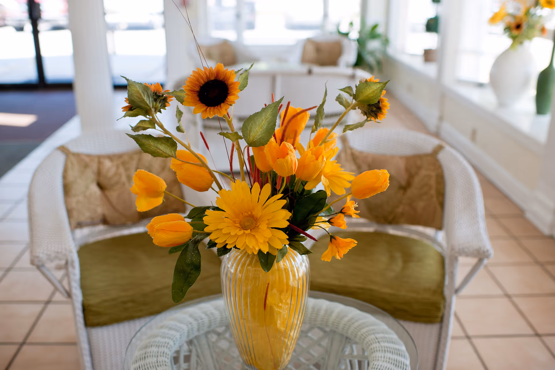 A close-up view of a glass table with a yellow vase holding a bouquet of artificial yellow and orange flowers, including sunflowers and tulips. Behind the table is a white wicker loveseat with green cushions and beige pillows, set in a bright room with tiled floors and large windows.