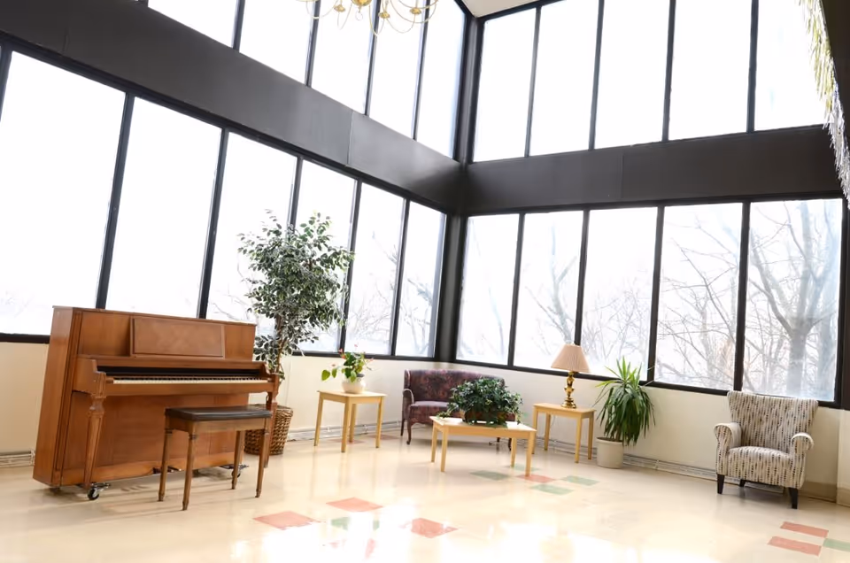 A bright and spacious living area with large floor-to-ceiling windows letting in natural light. The room features a wooden upright piano with a bench, several potted plants, a small sofa, an armchair, two side tables with a lamp, and a coffee table with a plant on it. The floor has a light-colored tile with some colored square patterns.