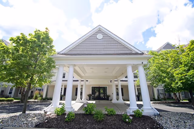 Covered portico entrance of a light-colored building with white columns, landscaping, and trees under a partly cloudy sky.