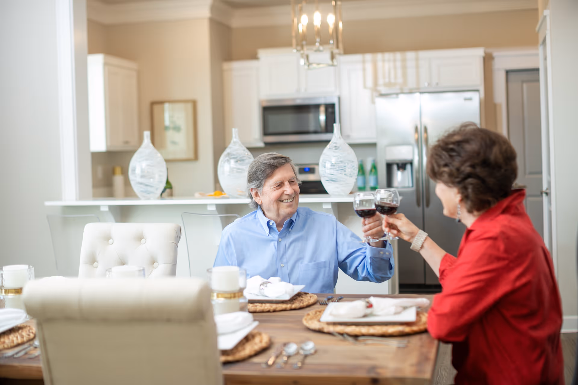 An elderly man and woman sitting at a dining table in a modern kitchen, smiling and clinking glasses of red wine in a toast. The kitchen features white cabinets, stainless steel appliances, and decorative glass vases on the counter.
