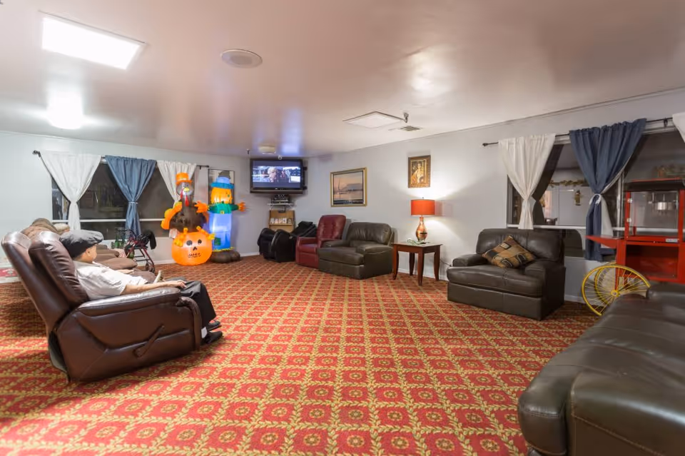 A spacious living room with patterned red carpet, several leather recliners and sofas arranged around the room, a wall-mounted TV playing a movie, and Halloween-themed inflatable decorations near the windows with blue and white curtains.