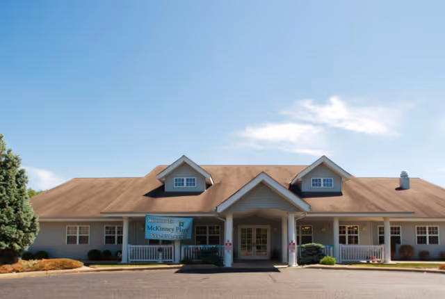 Front exterior view of a single-story building with a brown roof and light-colored siding under a clear blue sky. The building has a covered entrance with two white columns and a sign on the left side that reads 'Welcome to McKinney Place'. There are small bushes and landscaping around the building.