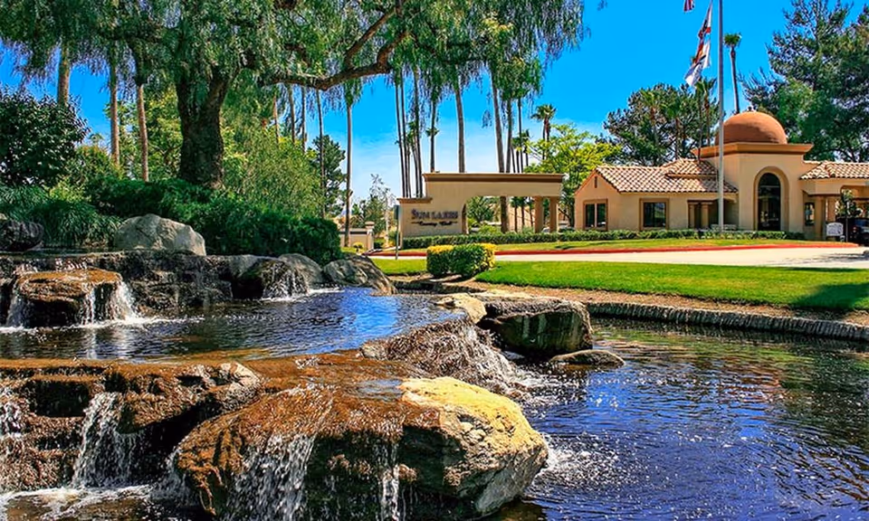 Rock-lined pond with small waterfalls in the foreground and the Sun Lakes Country Club entrance building and palm trees in the background.