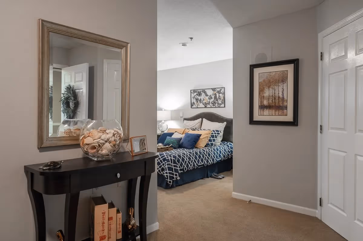 View of a hallway leading to a bedroom with a bed covered in blue and white patterned bedding and multiple pillows. A black console table with decorative items, including a glass bowl filled with seashells, is positioned against the wall under a large mirror. A framed artwork hangs on the wall near a closed white door.