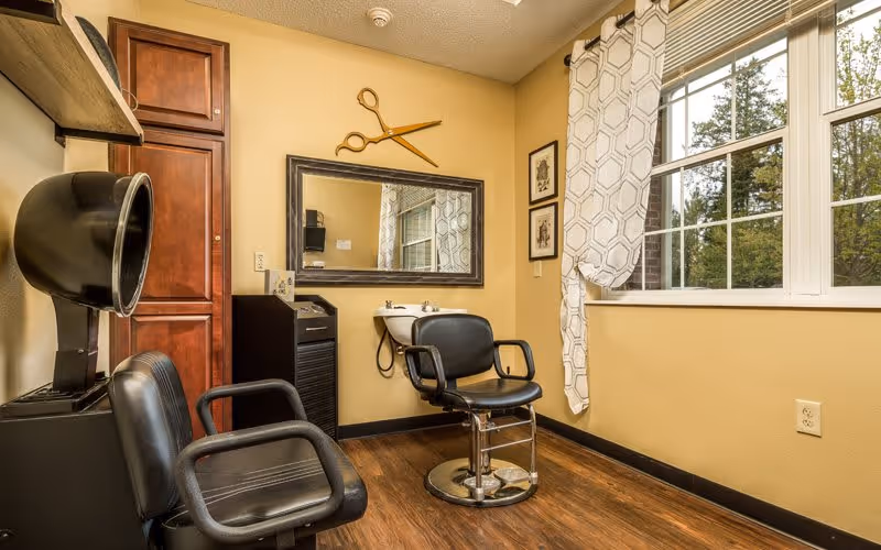Interior view of a small hair salon area with two black salon chairs, a hair dryer, a large mirror on the wall, a small sink, and a window with patterned curtains letting in natural light. There is a decorative large pair of scissors mounted above the mirror and wooden flooring.