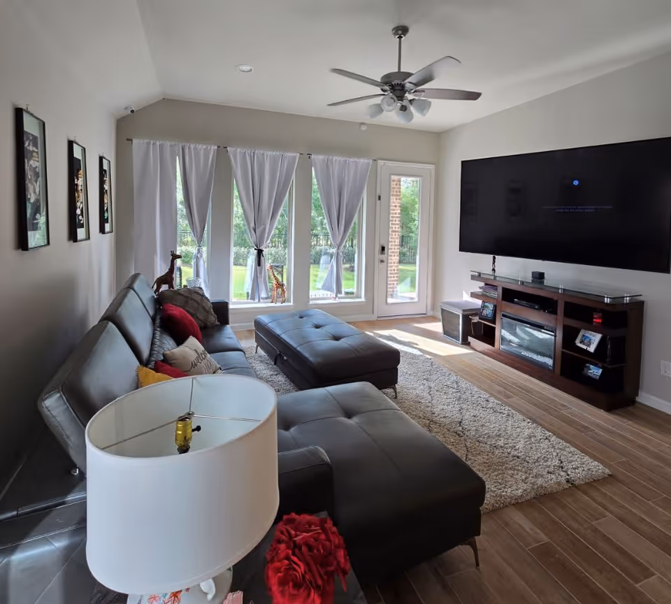 Bright living room with a leather sectional and ottoman, large wall-mounted TV, TV console, and tall windows with white curtains overlooking a yard.