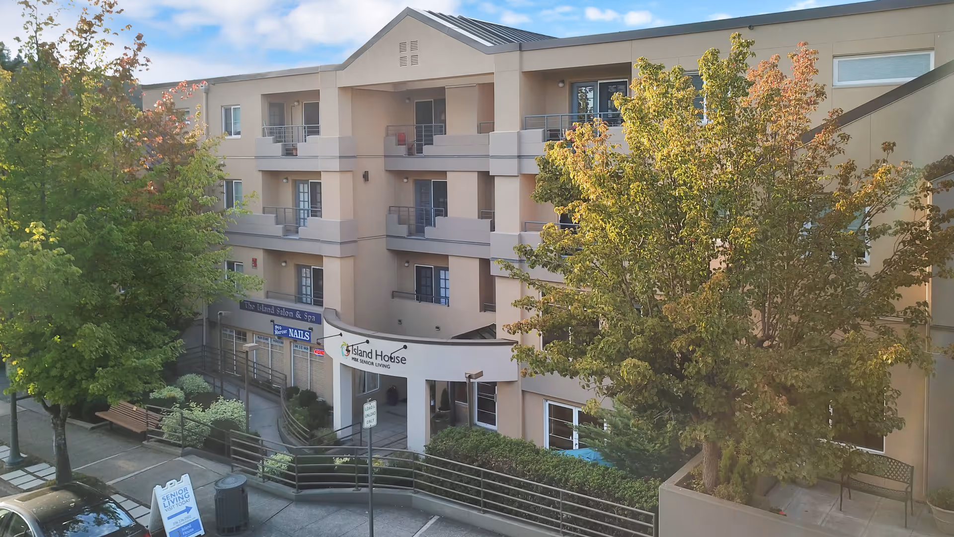 Exterior view of Island House Assisted Living facility showing a multi-story building with balconies, surrounded by trees and greenery. There is a sign for Island House and a sidewalk with benches and a trash can in front of the building.