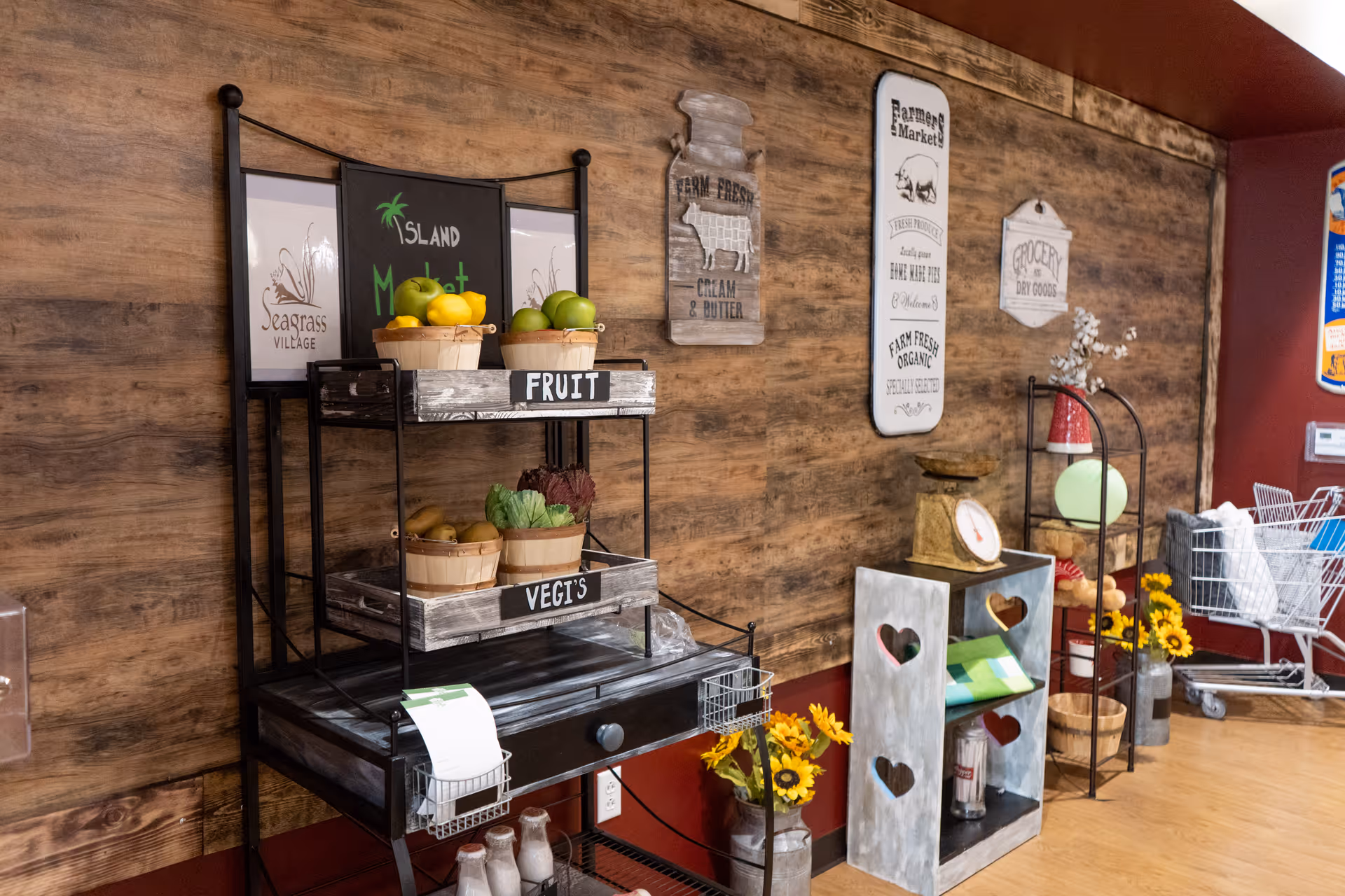 Interior view of a rustic-themed market display area with wooden walls. The display includes baskets labeled 'FRUIT' and 'VEGI'S' containing apples, lemons, and vegetables. Decorative signs on the wall read 'Farm Fresh Cream & Butter', 'Farmers Market', and 'Grocery Dry Goods'. There are shelves with sunflowers, a vintage scale, and a shopping cart in the background.