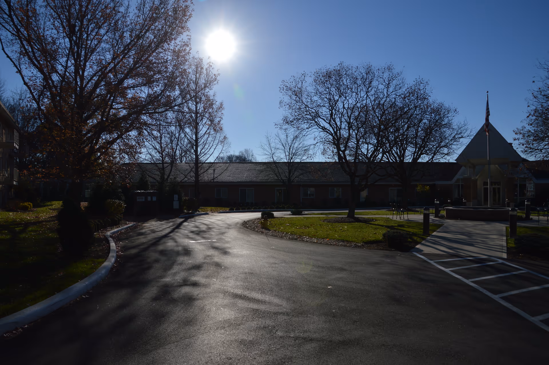 Entrance driveway and front facade of a single-story senior living building with trees, a flagpole, and the sun overhead.