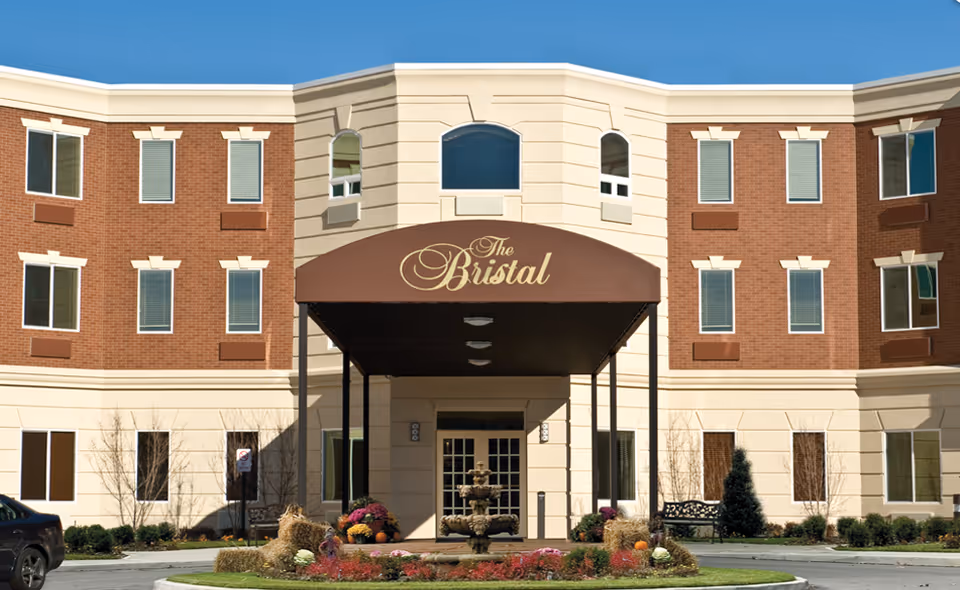 Front exterior view of The Bristal Assisted Living at East Northport building with a brown awning displaying the name The Bristal, a three-story structure with brick and beige facade, multiple windows, a fountain, and landscaped garden with flowers and pumpkins in front.