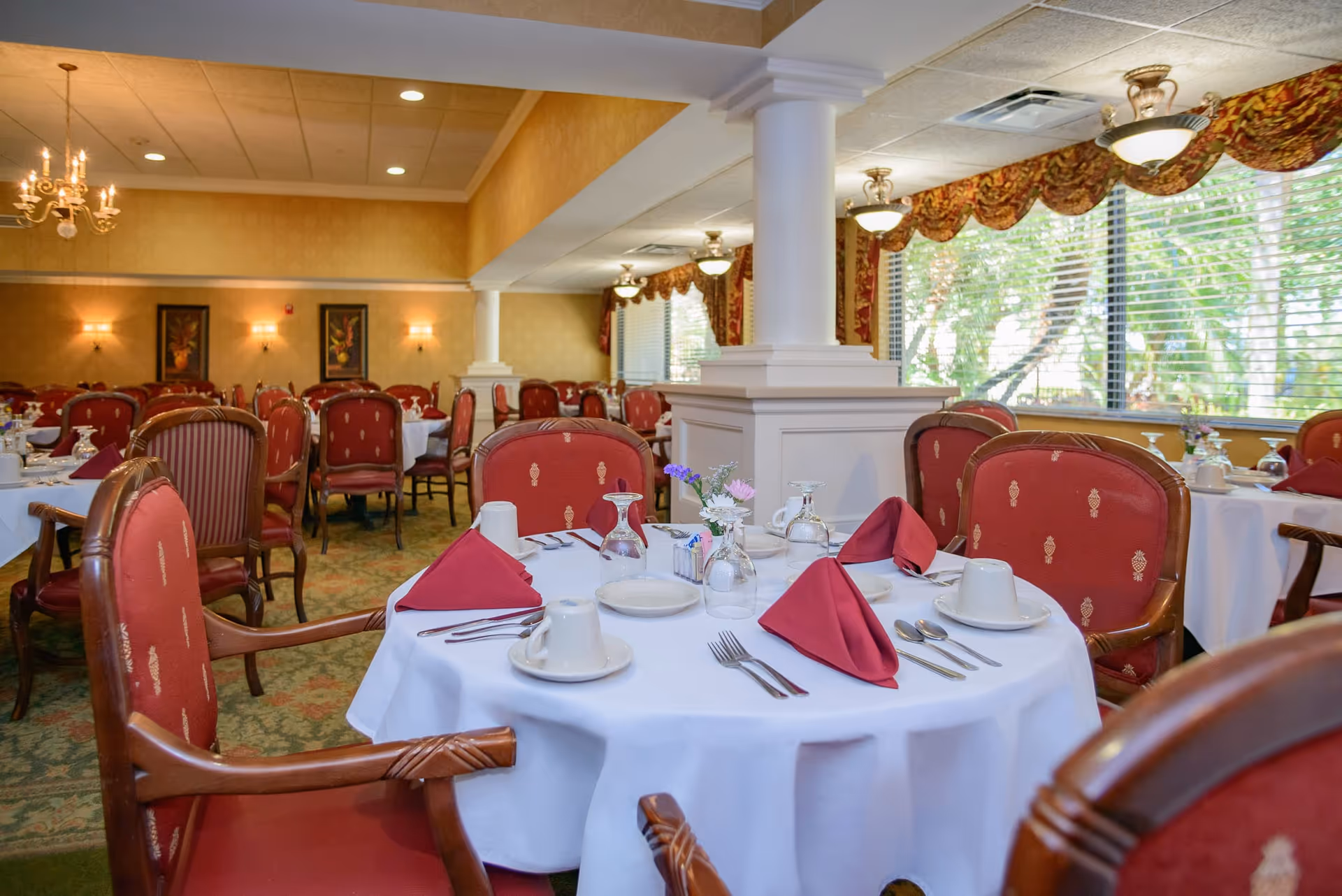 A dining room with round tables covered in white tablecloths, set with red folded napkins, white cups, plates, and silverware. The room features red upholstered chairs with wooden arms, large windows with patterned valances, chandeliers, and warm yellow walls with framed artwork.