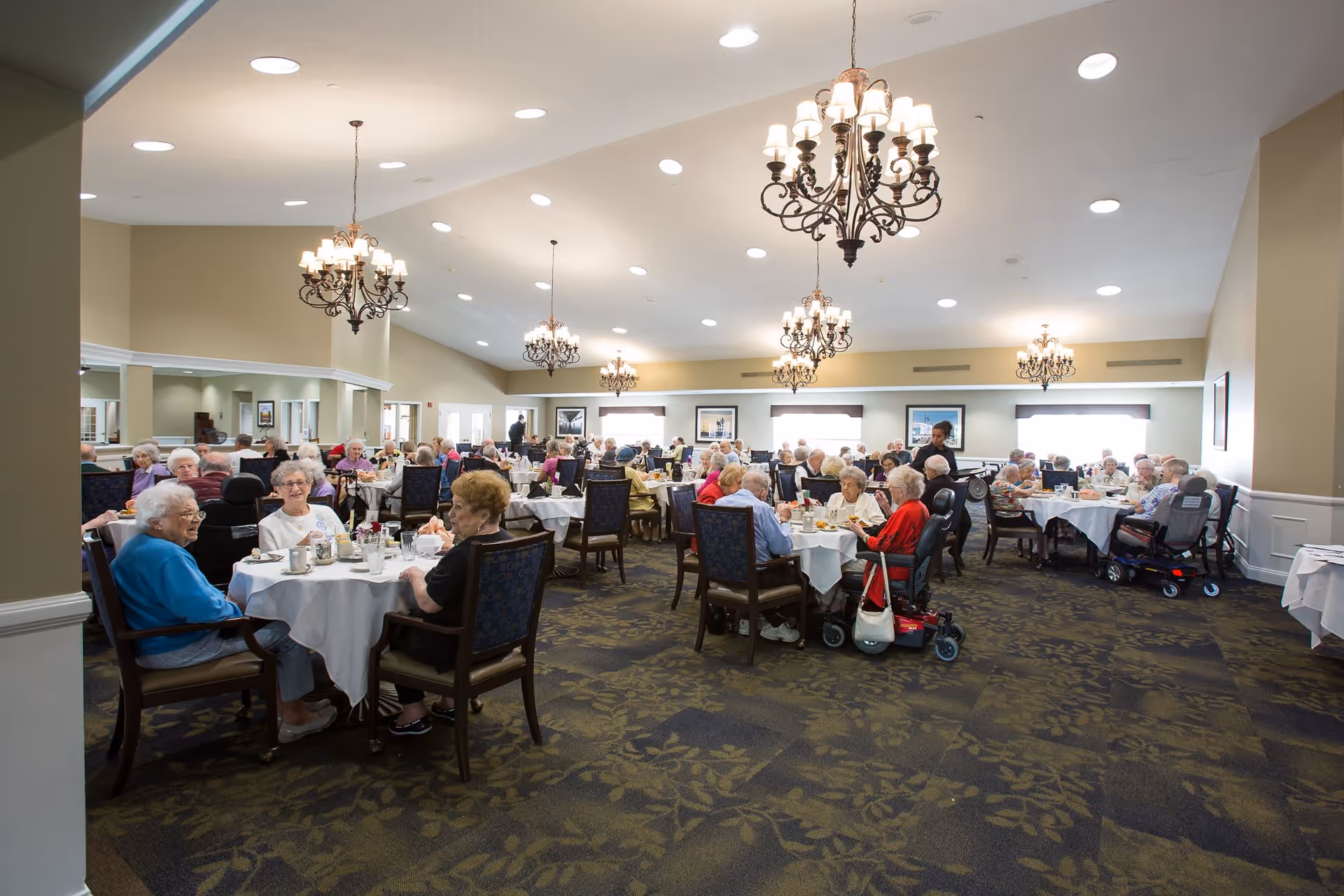A large dining room filled with elderly residents sitting at round tables covered with white tablecloths, enjoying a meal together. The room is well-lit with multiple chandeliers hanging from the ceiling and has a carpeted floor with a floral pattern.