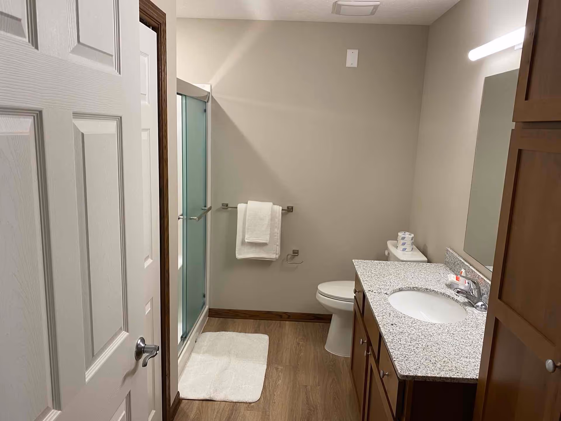 A clean bathroom with a white door partially open, a glass-enclosed shower with a white towel hanging on a towel rack, a white toilet with toilet paper on top, a granite countertop with a sink, faucet, and a mirror above it, and wooden cabinets below the countertop. The floor is wood-style and there is a white bath mat in front of the shower.