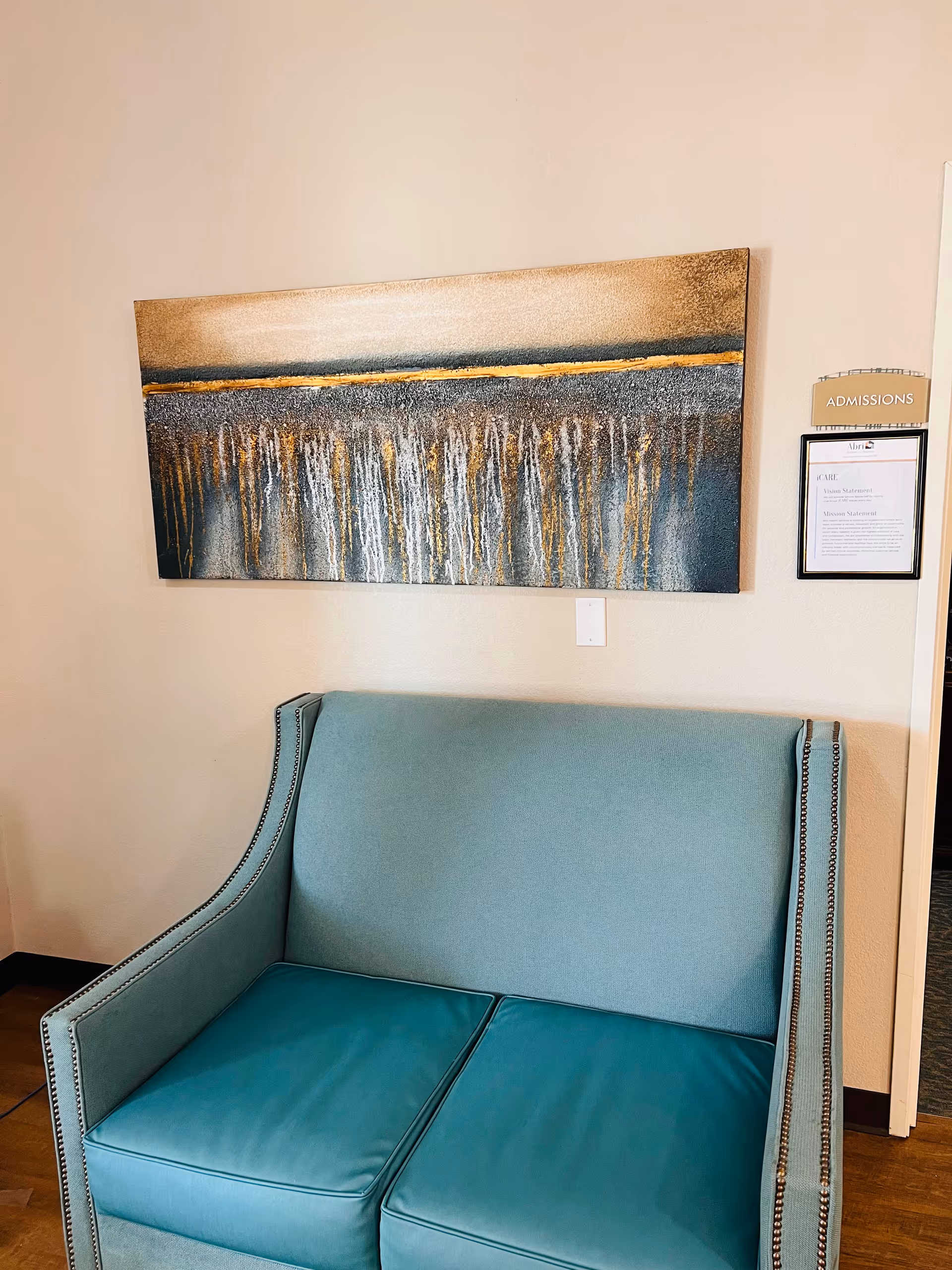 A teal upholstered loveseat with nailhead trim is positioned against a beige wall. Above the loveseat hangs a large abstract painting featuring vertical streaks in shades of gold, gray, and white. To the right of the painting is a small sign labeled 'ADMISSIONS' and a framed document beneath it.
