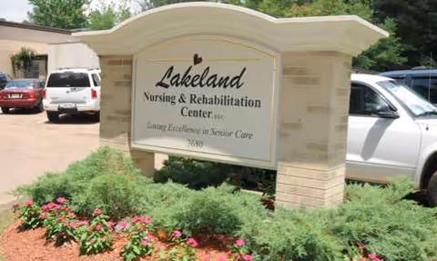Exterior view of Lakeland Nursing & Rehabilitation Center sign amid shrubs with parked cars nearby.