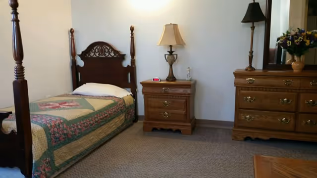 A traditional single bedroom with a wooden four-poster bed, matching nightstand and dresser, lamps, and a floral quilt.