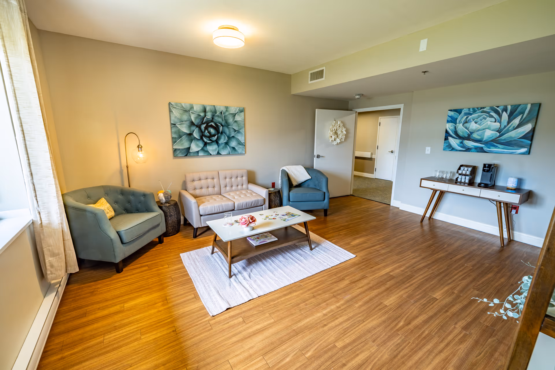 Bright furnished living room with two armchairs, a loveseat, a coffee table on a rug, and large floral wall art.
