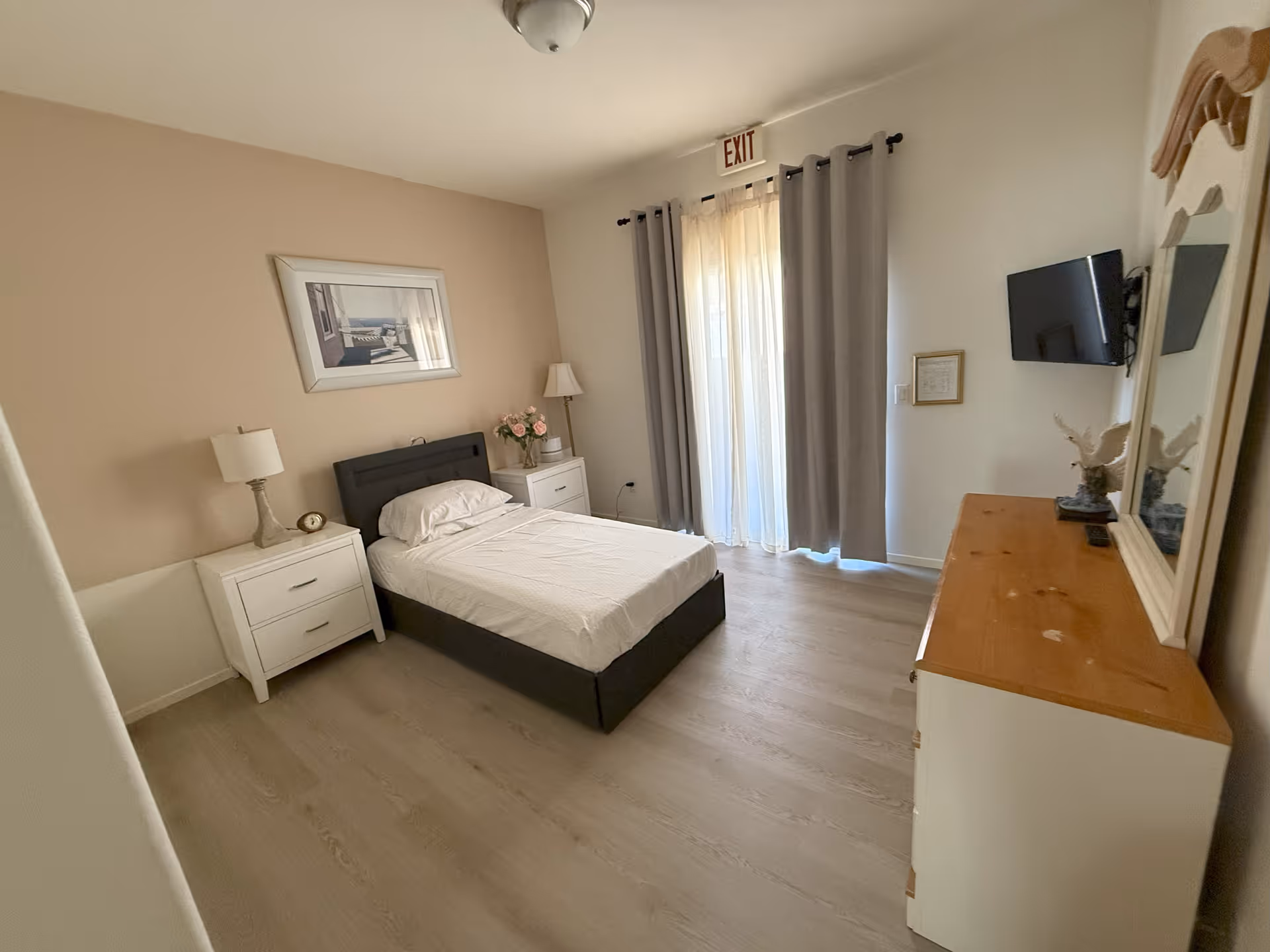 A simple, clean bedroom with a single bed centered against a beige wall. The bed has white bedding and is flanked by two white nightstands, each with a lamp. There is a framed picture hanging above the bed. A dresser with a wooden top and a large mirror is on the right side of the room, with a mounted flat-screen TV above it. Light-colored curtains cover a glass door with an exit sign above it. The floor is light wood.
