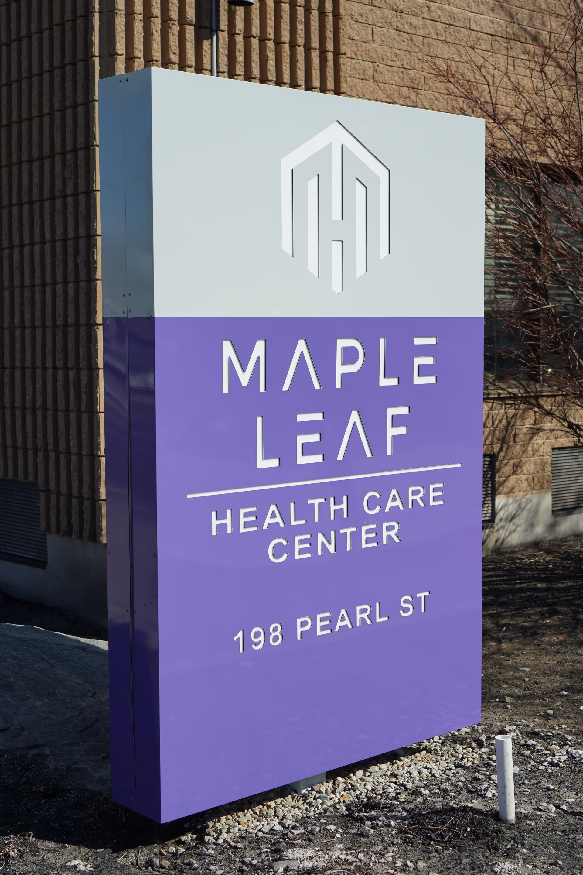 A tall rectangular sign outside a building with a white top and purple bottom. The sign displays the logo and name 'Maple Leaf Health Care Center' along with the address '198 Pearl St'. The background shows part of a brick building and some bare branches.