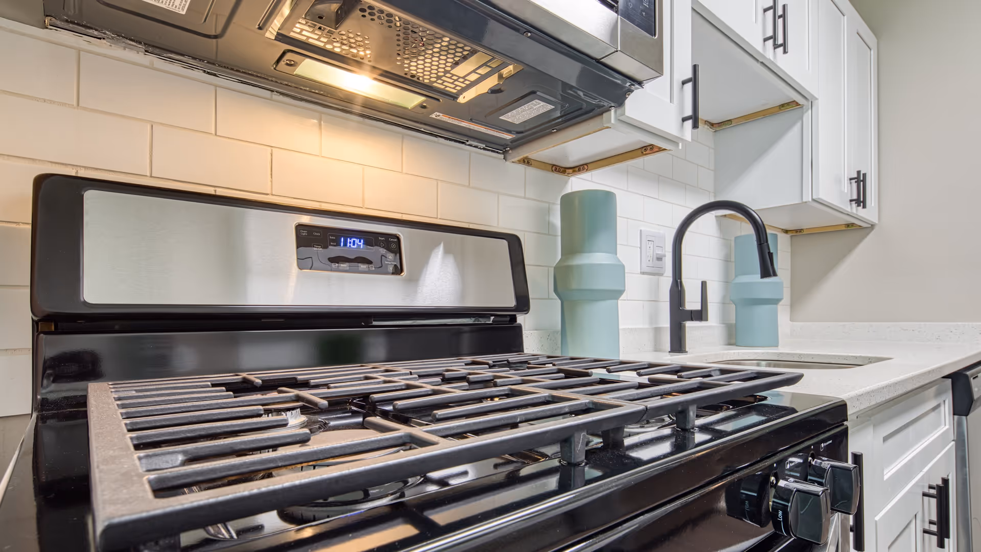 Close-up view of a modern kitchen stove with a stainless steel oven and black gas burners. Above the stove is a microwave with a light on. To the right, there is a white countertop with a black faucet and two decorative blue-green vases. White cabinets with black handles are visible above and below the countertop.