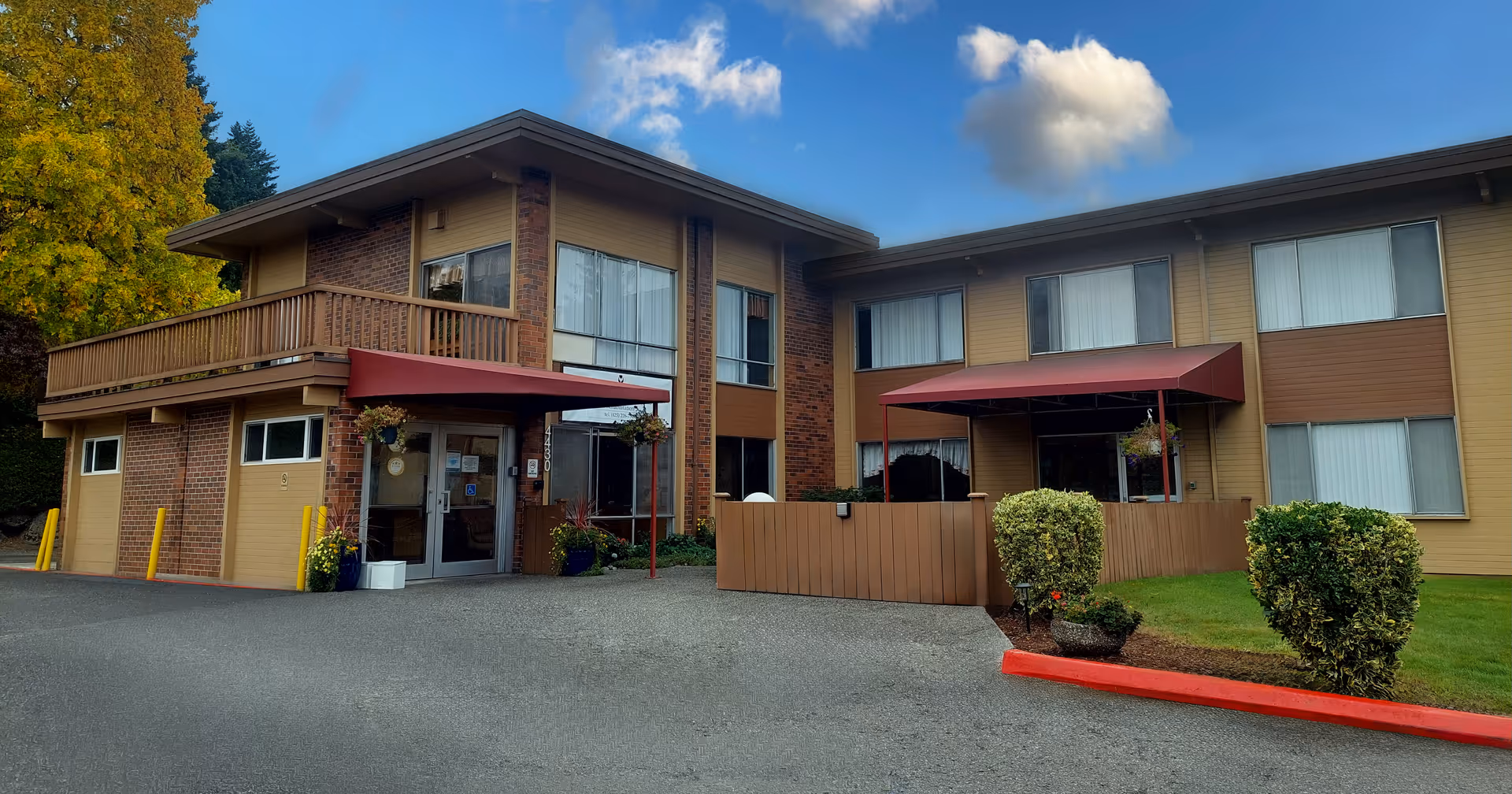 Two-story brick and wood nursing facility front with red awnings, an entrance driveway, and trimmed landscaping under a blue sky.