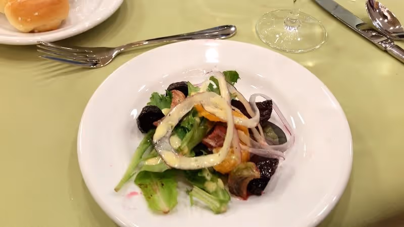 A small salad on a white plate with mixed greens, sliced onions, and other vegetables, drizzled with dressing. A fork is placed above the plate, a bread roll on a small plate is partially visible to the left, and a wine glass and silverware are on the right side on a light green tablecloth.