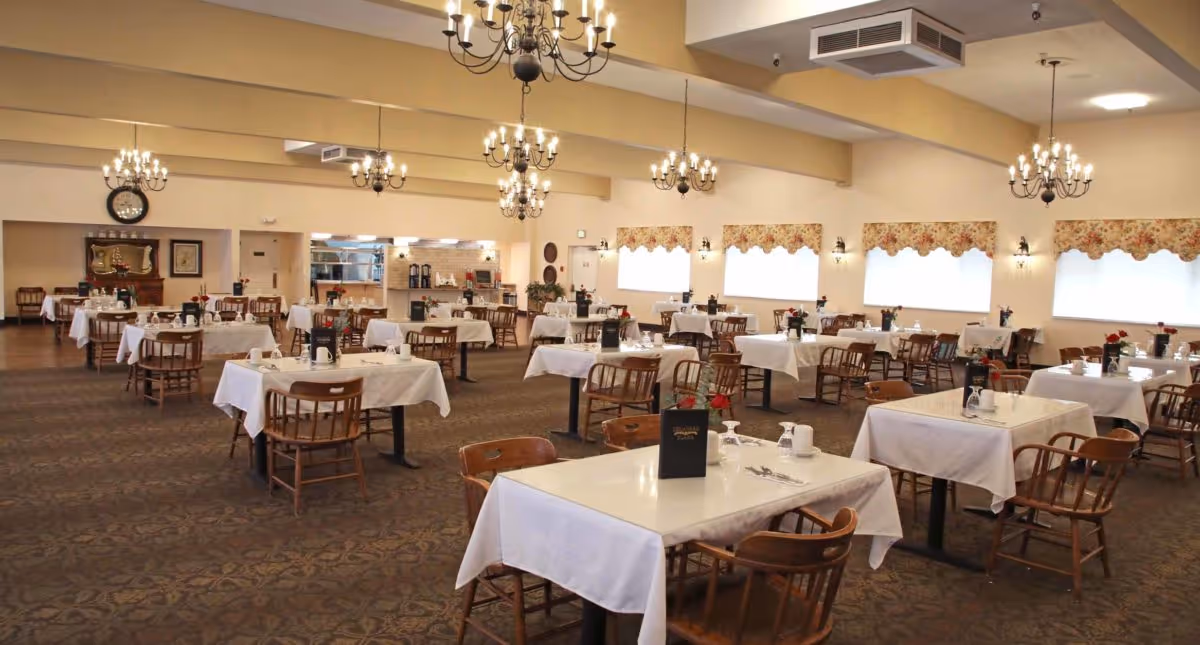 Spacious dining room with multiple tables covered in white tablecloths, each set with cups, utensils, and small flower vases. Wooden chairs surround the tables, and chandeliers hang from the ceiling. The room has large windows with floral valances and a carpeted floor.