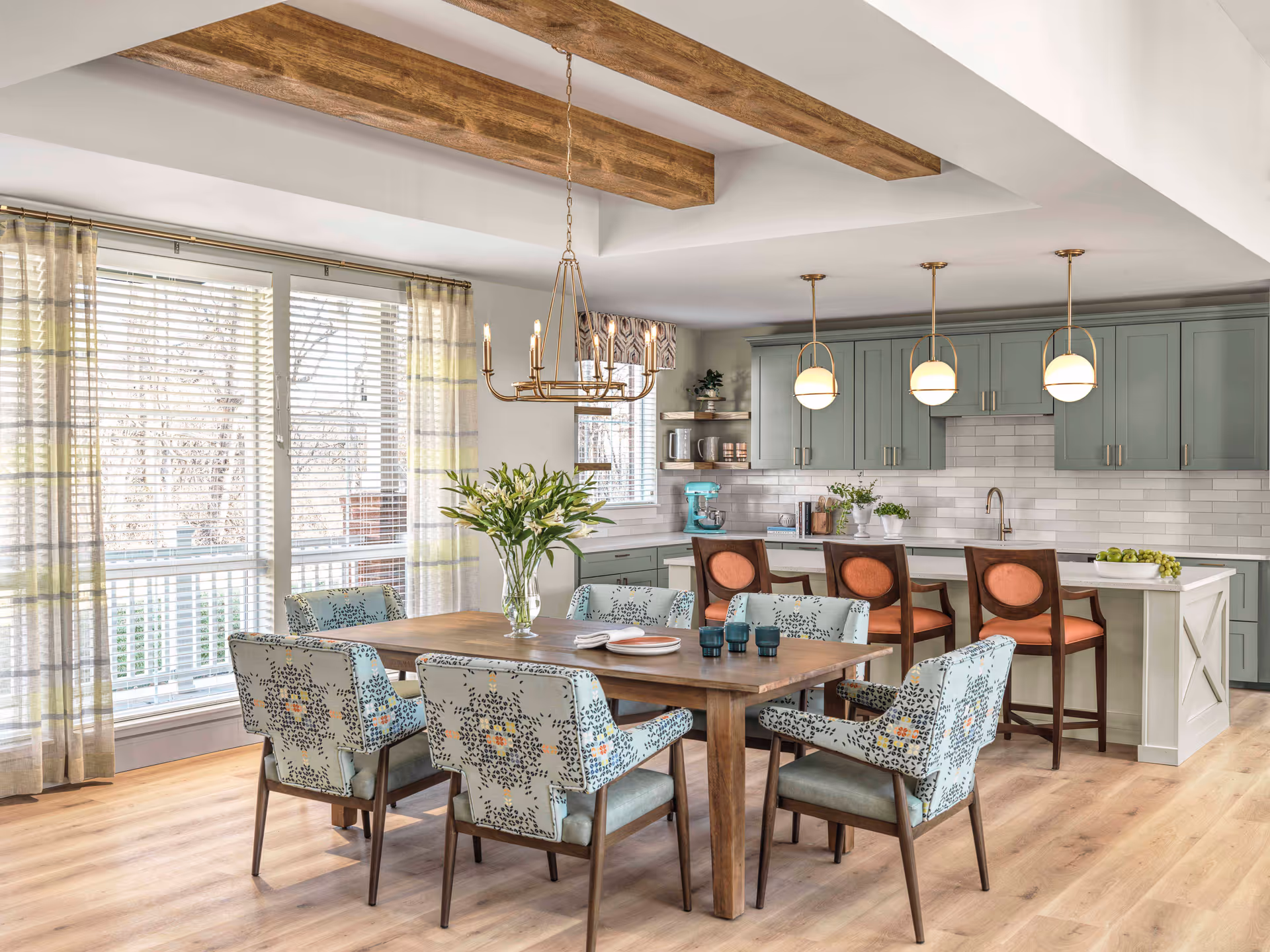 A bright and modern dining area with a wooden dining table surrounded by six upholstered chairs with patterned fabric. Behind the dining table is a kitchen with light green cabinets, a white countertop island with three orange cushioned bar stools, and three pendant lights hanging above the island. Large windows with sheer curtains allow natural light to fill the space, and wooden ceiling beams add warmth to the room.