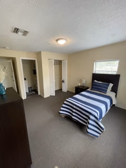 A simple bedroom with a single bed covered in a blue and white striped blanket and matching pillow. There is a small nightstand with a lamp next to the bed, a window with blinds above the headboard, an open closet with white bi-fold doors, and a dark dresser with a decorative vase on top. The room has beige walls and carpeted floor.