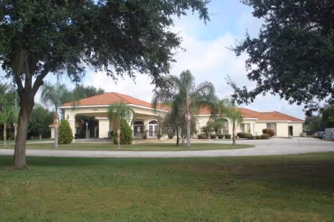 Single-story building with a red-tiled roof surrounded by palm trees and other greenery under a partly cloudy sky, viewed from a grassy area with a circular driveway in front.