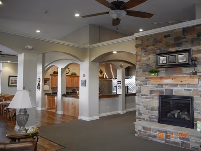 Interior view of a senior living facility showing a cozy common area with a stone fireplace on the right, wooden ceiling fan above, and an open kitchen area with wooden cabinets and stainless steel appliances in the background. There is a dining area visible to the left with tables and chairs, and the flooring transitions from carpet to wood.