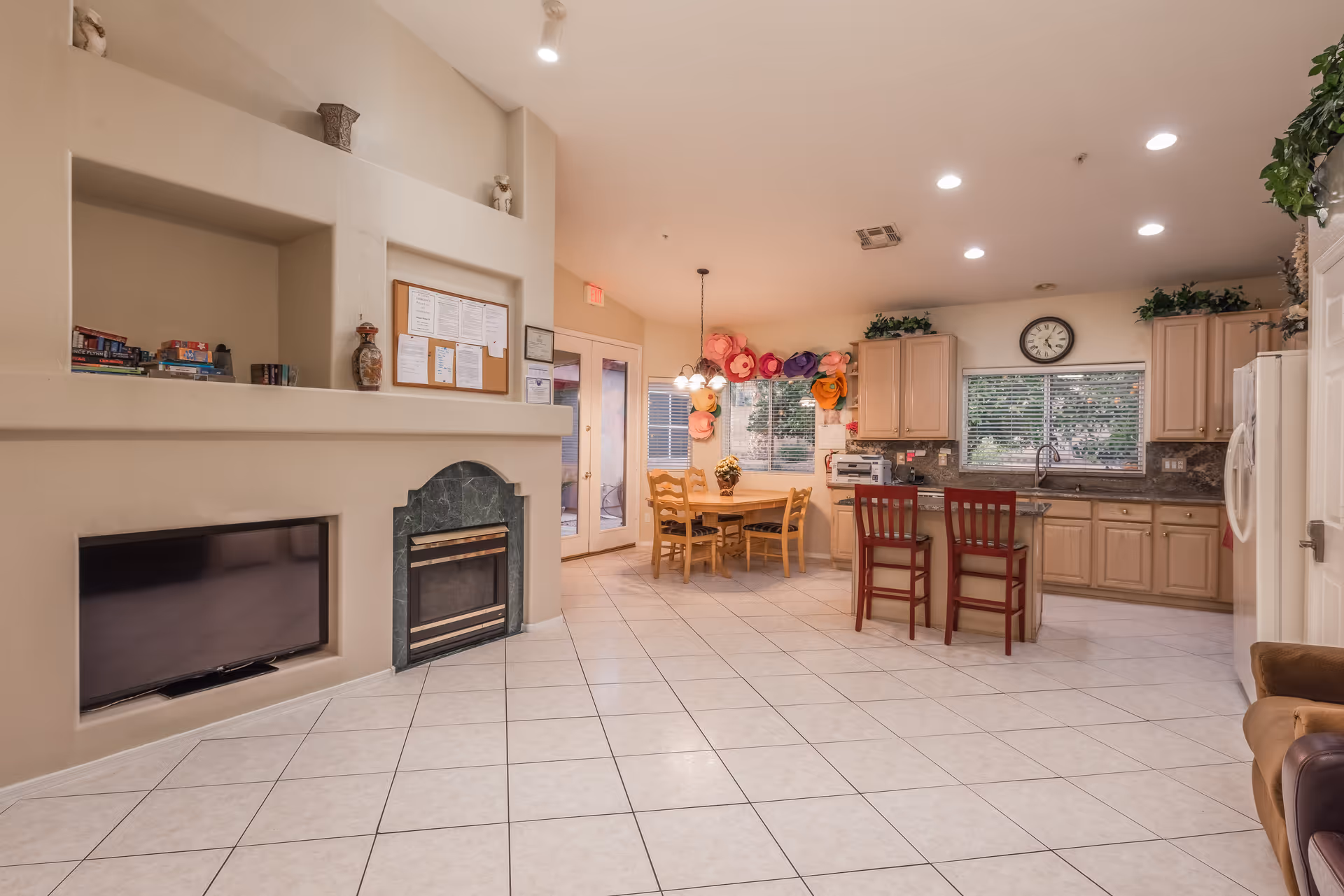 Open common area with a kitchen, dining table and chairs, fireplace, and seating in a senior living facility.