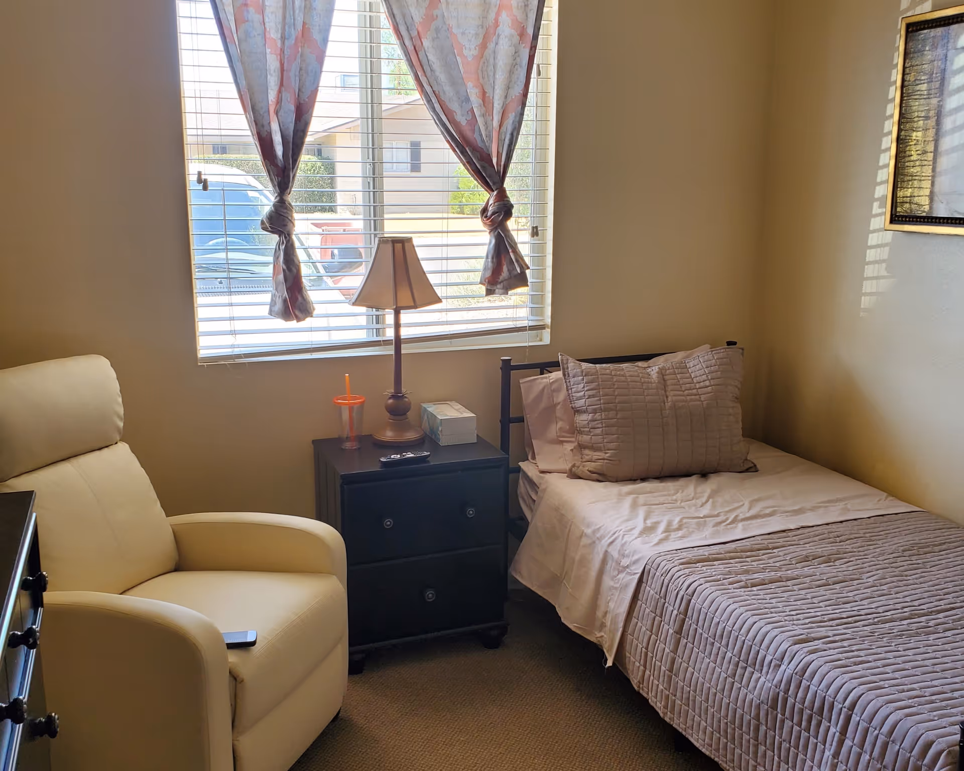 Small furnished bedroom with a single bed, nightstand and lamp, a reclining armchair, and a window with tied curtains.
