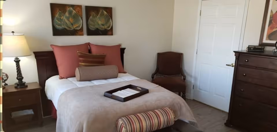 A cozy bedroom with a neatly made bed featuring pink and brown pillows, a beige blanket, and a tray on top. There is a wooden nightstand with a lamp on the left side of the bed, a brown upholstered chair in the corner, and a dark wooden dresser on the right. Two leaf-themed paintings hang above the bed, and a white door is visible in the background.