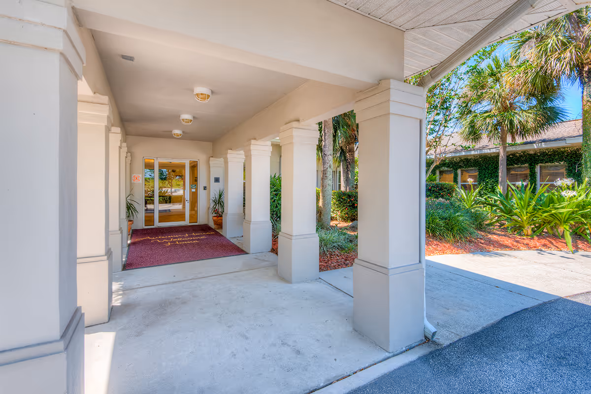 Covered entrance area of a building with white columns and a maroon welcome mat leading to glass double doors. There are potted plants on either side of the doors and greenery with palm trees visible outside.