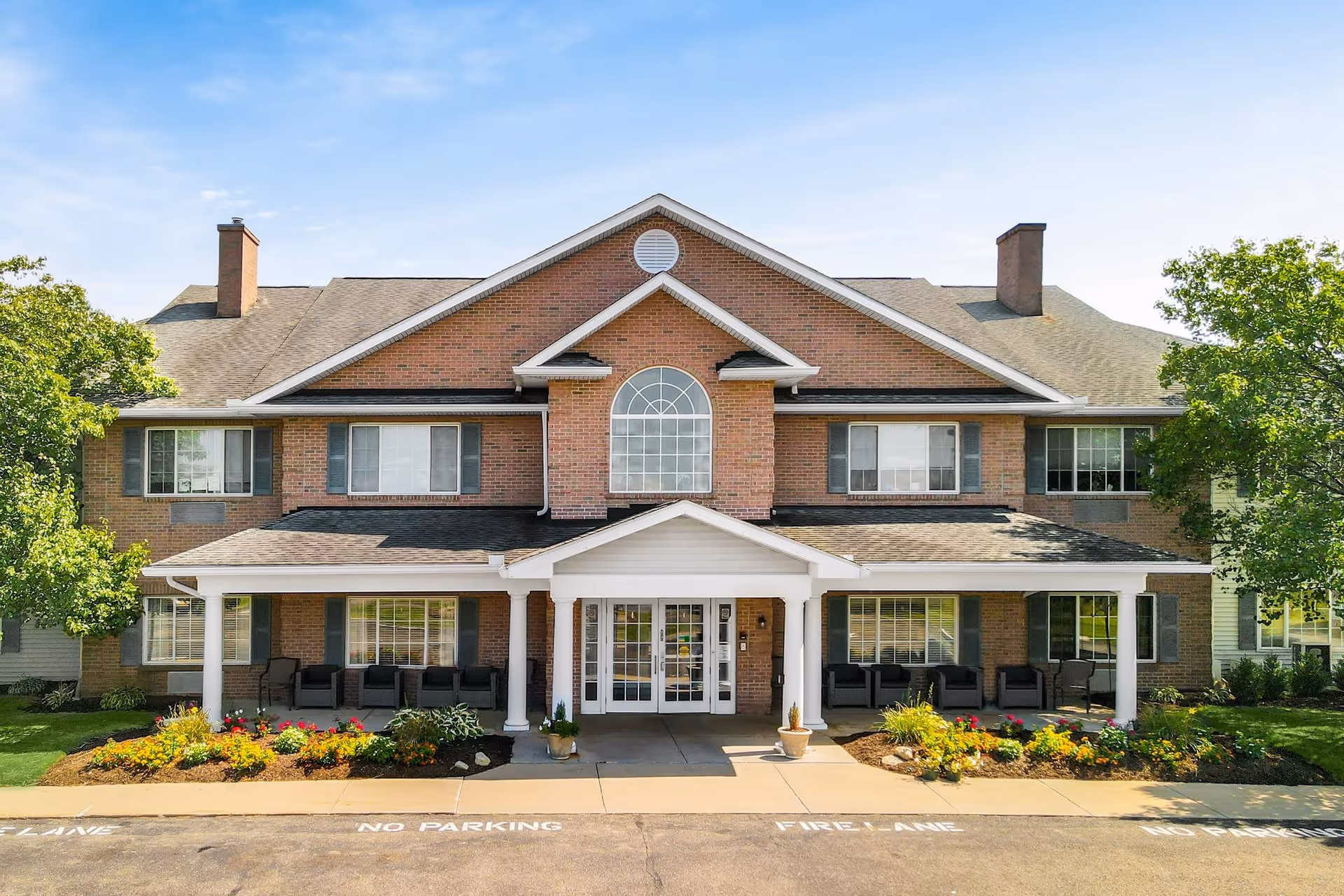 Front exterior view of a two-story brick building with a covered entrance supported by white columns. The building has multiple windows with shutters, two chimneys, and a landscaped area with flowers and shrubs in front. There are chairs arranged on the porch under the covered entrance. The sky is clear and blue.
