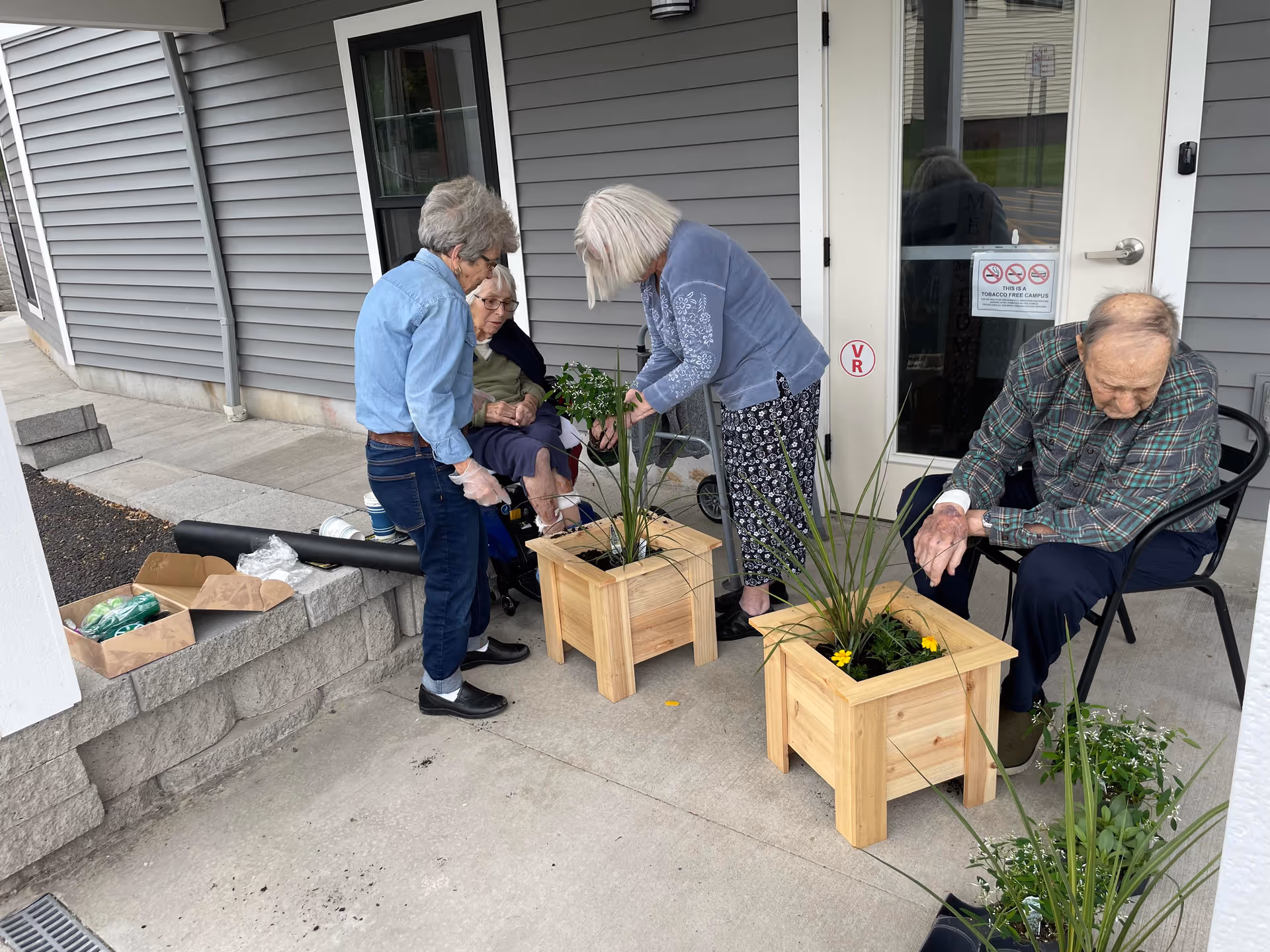 Four older adults planting flowers in wooden planters on the building's front patio near the entrance.