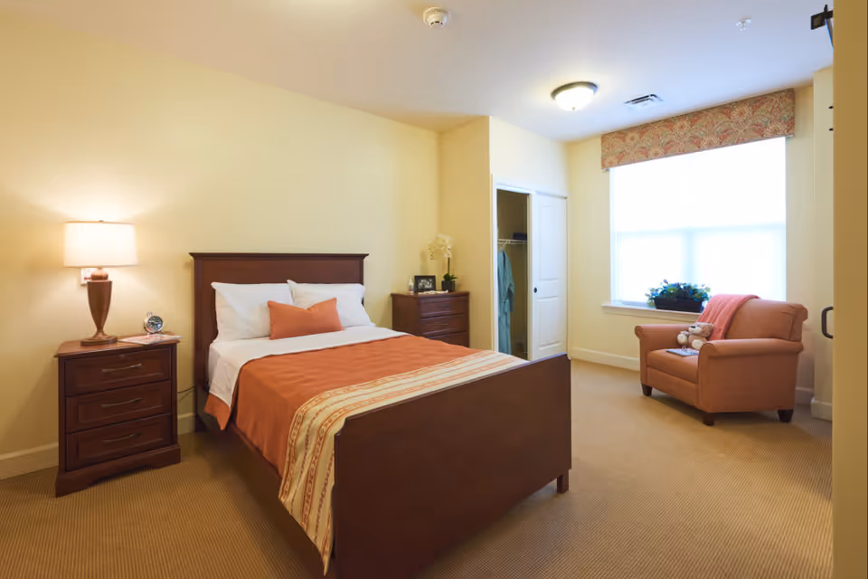 A cozy bedroom in an assisted living facility featuring a wooden bed with white and orange bedding, a nightstand with a lamp and clock, a dresser, an open closet with hanging clothes, and an armchair with a teddy bear near a large window with a decorative valance.