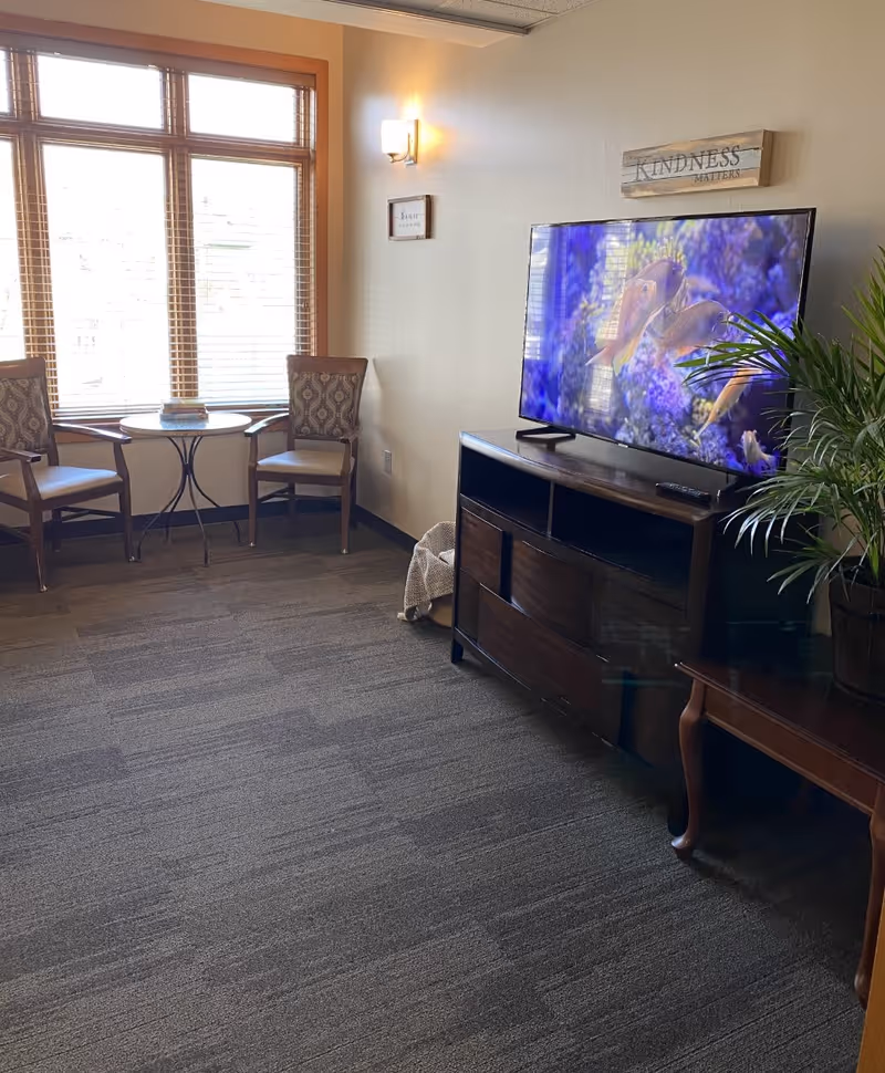 A cozy living room area with two patterned chairs and a small round table near a large window with wooden blinds. A flat-screen TV displaying an underwater scene with fish is placed on a dark wooden TV stand. A green potted plant is on a small wooden side table next to the TV stand. The wall has two framed signs, one reading 'KINDNESS MATTERS'.