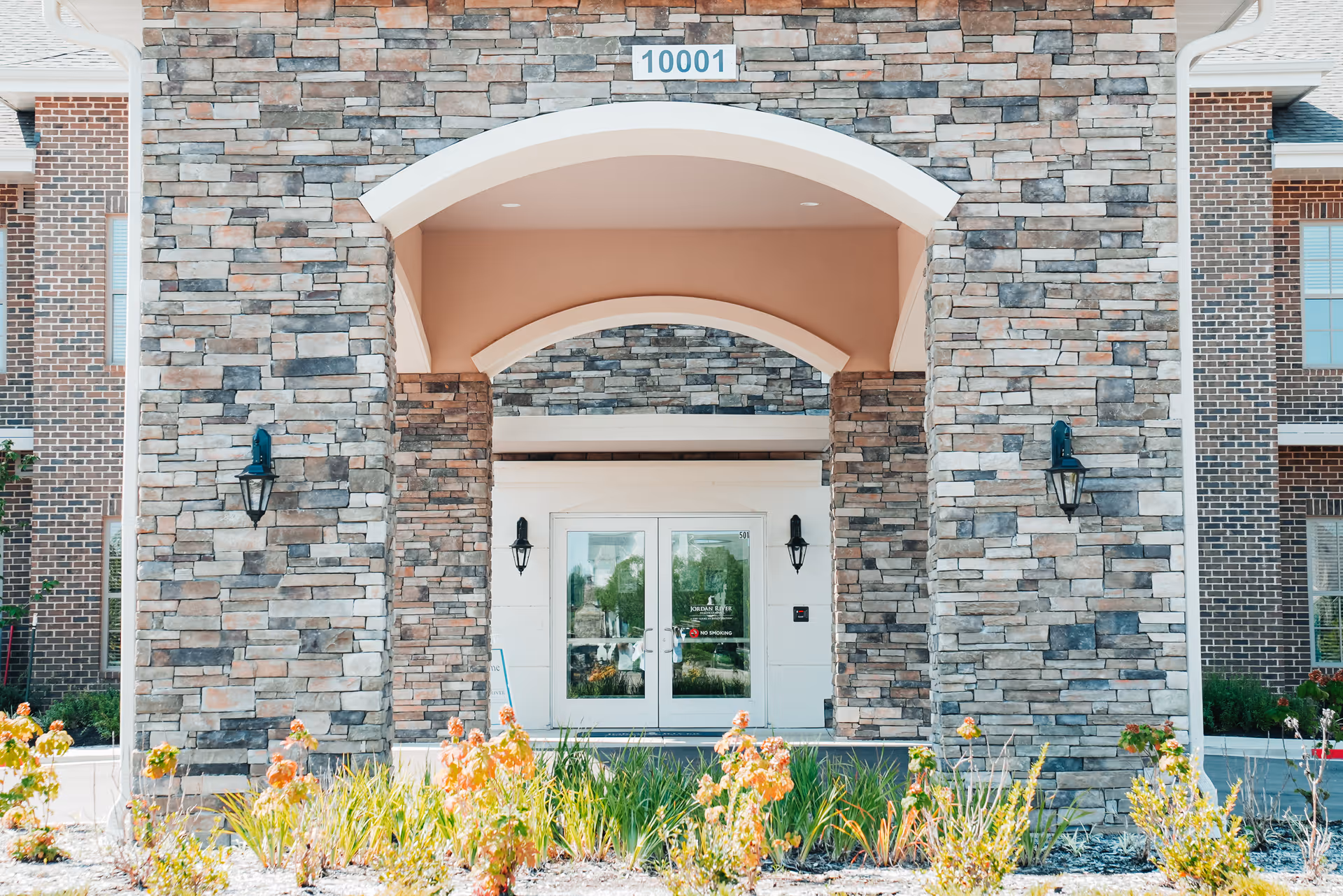 Entrance of a building with stone and brick exterior walls, featuring a large archway and double glass doors. The address number 10001 is displayed above the arch. There are outdoor wall lanterns on either side of the entrance and some landscaping with plants and flowers in front.