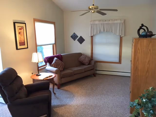 A cozy living room with a brown sofa adorned with patterned pillows and a purple throw blanket, a dark recliner chair, a wooden side table with a lamp, two windows with white blinds and a valance, a ceiling fan, framed wall art, a wooden cabinet with decorative items on top, and a green plant in the corner.