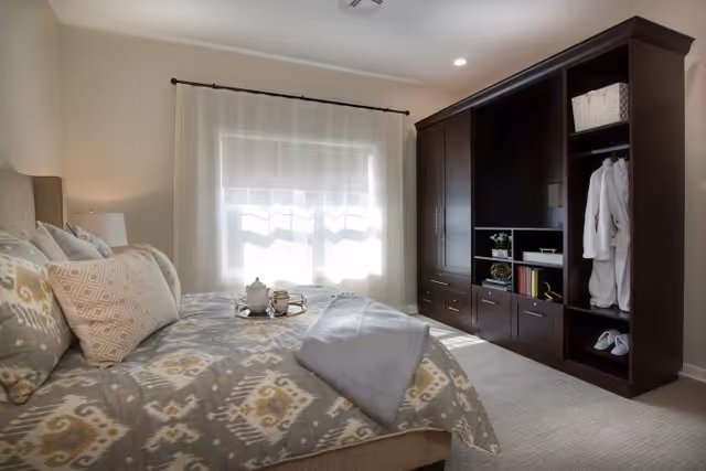 A cozy bedroom with a bed covered in patterned bedding and multiple pillows. A tray with a teapot and cups is placed on the bed. There is a large window with sheer white curtains letting in natural light. On the right side, there is a dark wooden wardrobe with open shelves containing books, baskets, robes, and slippers. The room has a soft carpeted floor and neutral-colored walls.