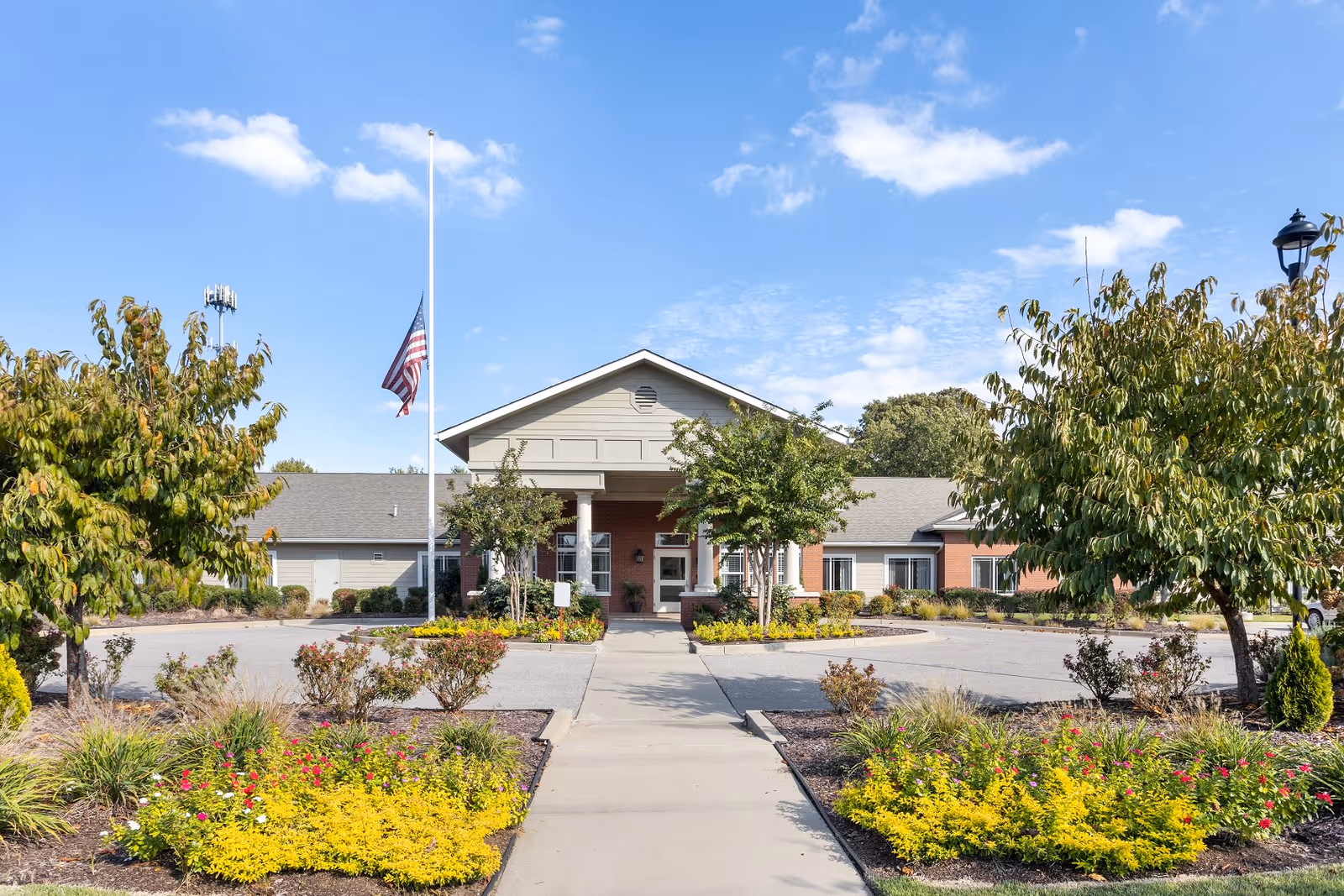 Front exterior view of Quail Ridge Assisted Living & Memory Care building with a walkway leading to the entrance, landscaped garden beds with yellow and red flowers, trees on both sides, and an American flag at half-mast against a blue sky with scattered clouds.