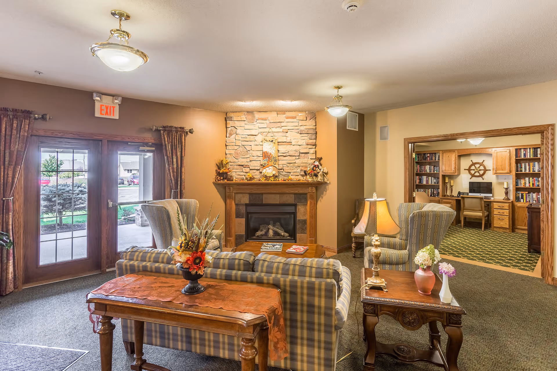 A cozy living room area in a senior living facility with plaid upholstered chairs and sofa arranged around a wooden coffee table in front of a stone fireplace decorated with autumn-themed ornaments. To the right, there is a wooden side table with a lamp and flower vases. In the background, an open doorway leads to a room with bookshelves, a desk, and a ship's wheel decoration on the wall. Double glass doors with curtains lead outside, and an exit sign is visible above the doors.